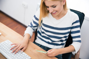 Happy young woman holding bank card and typing on keyboard in office